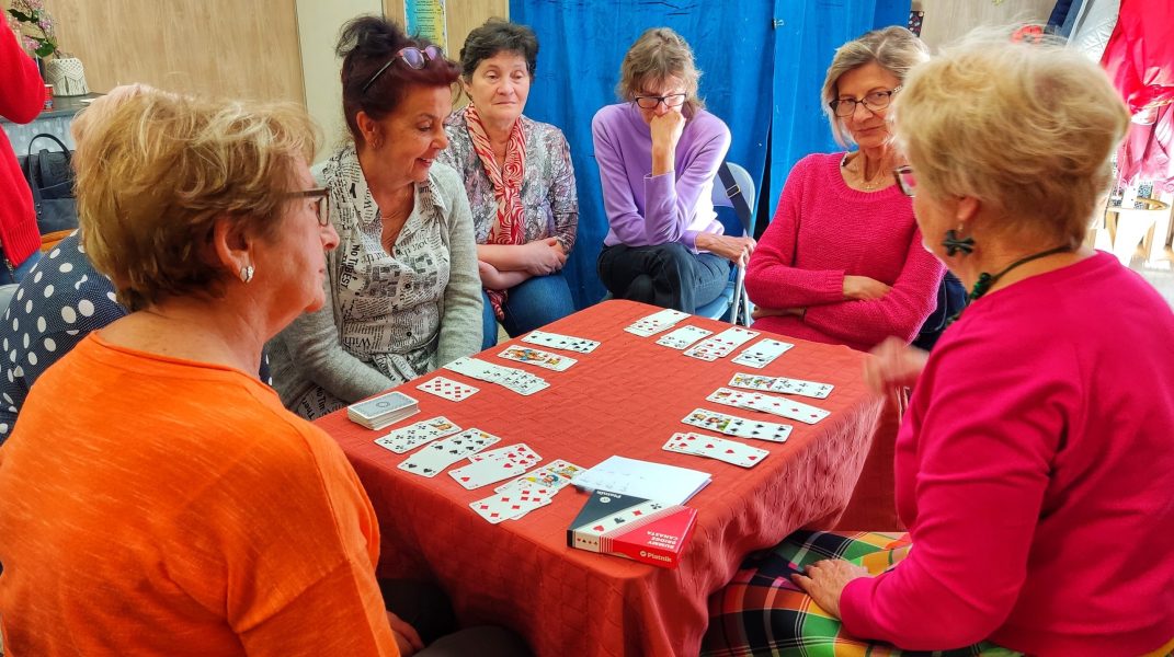 Sześć kobiet siedzi przy stole przykrytym czerwonym obrusem, na którym rozłożone są karty do gry w brydża. Uczestniczki uważnie obserwują sytuację na stole, a jedna z nich, ubrana w różową bluzkę, trzyma karty w dłoniach