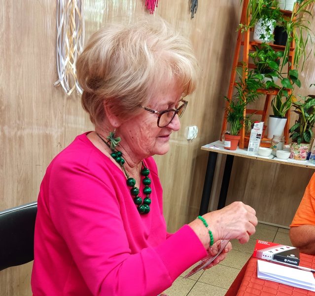 Seniorka w okularach, różowej bluzce i zielonych koralach siedzi przy stole, trzymając w dłoniach karty do gry w brydża. W tle widoczna jest ściana z makramami oraz regał z roślinami doniczkowymi, takimi jak zielistka i sansewieria