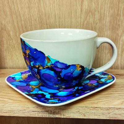 Biała ceramiczna filiżanka i kwadratowy spodek udekorowane abstrakcyjnym wzorem wykonanym tuszami alkoholowymi w odcieniach intensywnego błękitu, turkusu i fioletu. Wzór przypomina płynne, przenikające się plamy barwne, które są dodatkowo ozdobione nieregularnymi akcentami złotego szlagmetalu. Zestaw stoi na jasnej, drewnianej półce na tle drewnianej ściany