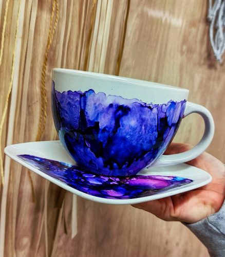 Ceramiczna biała filiżanka i spodek z abstrakcyjnym zdobieniem wykonanym tuszami alkoholowymi w intensywnych odcieniach fioletu, granatu i indygo. Wzór na naczyniu tworzy malarskie, przenikające się plamy o nieregularnych krawędziach, przypominające strukturę minerałów. Zestaw trzymany jest w dłoni na tle drewnianej ściany