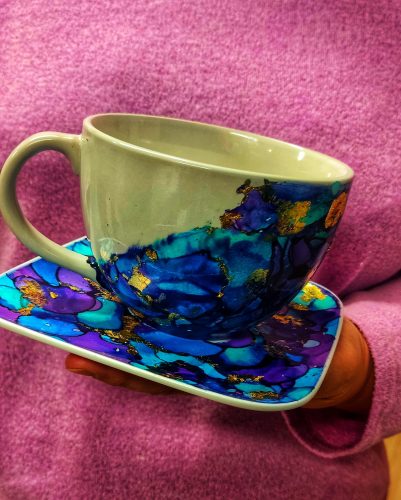 Biała ceramiczna filiżanka ze spodkiem, trzymana na dłoni na tle różowego swetra. Naczynia ozdobione są tuszami alkoholowymi w odcieniach głębokiego błękitu, turkusu i fioletu, z licznymi płatkami złotego szlagmetalu wtopionymi w barwną kompozycję