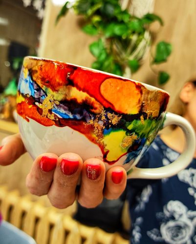 Zbliżenie na dłoń z czerwonym manicure trzymającą białą ceramiczną filiżankę ozdobioną tuszami alkoholowymi. Wzór na naczyniu tworzy dynamiczną, wielobarwną kompozycję w odcieniach czerwieni, pomarańczu, żółci, zieleni i błękitu, z licznymi płatkami złotego szlagmetalu. W tle widoczna jest rozmyta postać i zielone liście rośliny