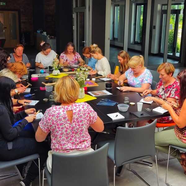 Grupa kobiet siedząca przy dużym, ciemnym stole w nowoczesnym, jasnym wnętrzu podczas warsztatów rękodzielniczych. Uczestniczki są skupione na precyzyjnej pracy manualnej, używając młotków i specjalistycznych narzędzi do tworzenia wzorów na białych bazach. Na stole znajdują się motki kolorowych sznurków, słoiczki z akcesoriami oraz dekoracyjne wazony z polnymi kwiatami