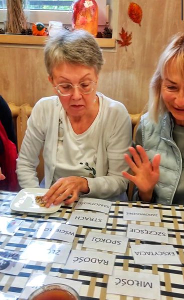 Dwie seniorki podczas warsztatów terapeutycznych w Warsztatowni. Na stole leżą karty z nazwami emocji w języku polskim, takimi jak: „Radość”, „Miłość”, „Spokój”, „Ekscytacja” oraz „Strach”