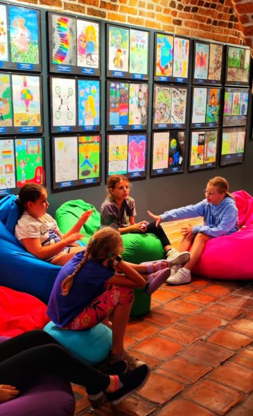 Czworo dzieci siedzi na kolorowych pufach sako (niebieskiej, zielonej i różowej) w sali o ceglanej podłodze, prowadząc ożywioną rozmowę. W tle ściana gęsto pokryta oprawionymi rysunkami dzieci przedstawiającymi różne postacie i sceny