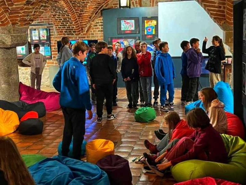 Grupa dzieci uczestniczy w warsztatach o emocjach w klimatycznej sali z ceglanymi łukami. Część dzieci siedzi na kolorowych pufach sako, a inne stoją w grupie, rozmawiając i integrując się przed ekranem i tablicą z pracami