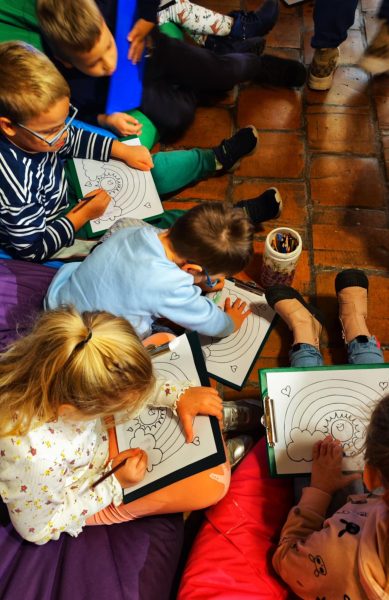 Widok z góry na grupę dzieci siedzących na podłodze, które kolorują kredkami rysunki słońca i tęczy umieszczone na podkładkach z klipsem. Obok stoi pojemnik z kolorowymi kredkami