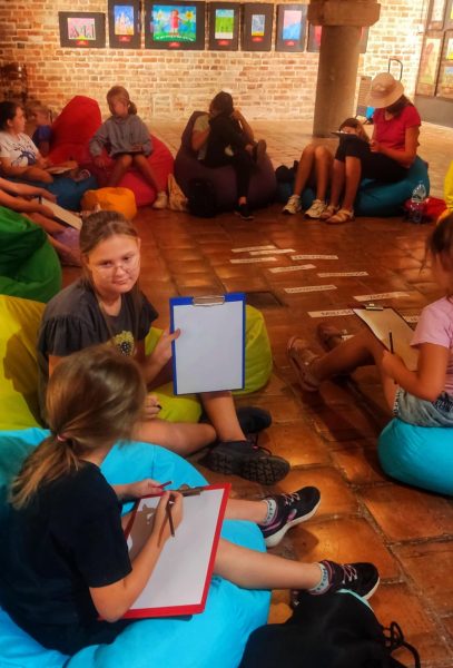 Grupa dzieci siedzi na kolorowych pufach sako ułożonych w krąg na ceglanej podłodze. W centralnym punkcie kręgu leżą białe paski z napisami określającymi emocje (m.in. smutek, strach, samotność, miłość). Niektóre dzieci trzymają podkładki z kartkami, przygotowując się do rysowania lub omawiania swoich prac