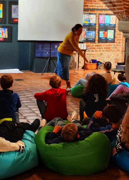 Grupa dzieci i młodzieży siedzi wygodnie na kolorowych pufach sako (turkusowych, zielonych, czerwonych) w sali z ceglanym sklepieniem łukowym. W tle widać ekran projekcyjny oraz prowadzącą zajęcia Moniki Wojdyło w żółtej bluzce i dżinsowej spódnicy