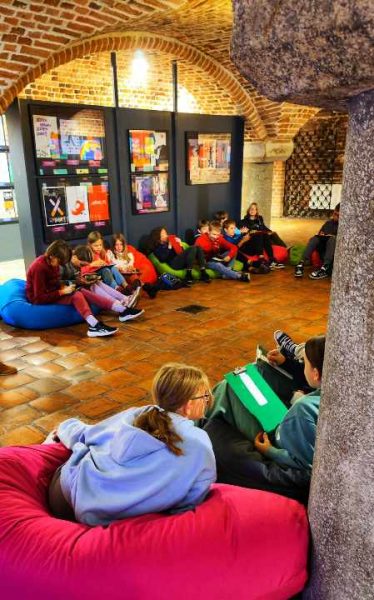 Grupa dzieci i młodzieży siedzi na kolorowych pufach sako (niebieskich, czerwonych, fioletowych) na ceglanej podłodze w sali o łukowym sklepieniu. W tle widać ściany z czerwonej cegły oraz galerię prac plastycznych