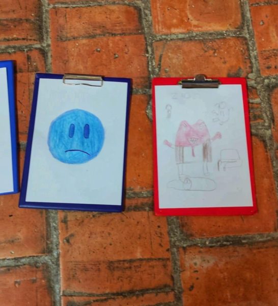 Dwie prace plastyczne na podkładkach z klipsem (niebieskiej i czerwonej) leżące na ceglanej podłodze. Pierwsza przedstawia dużą, smutną niebieską buźkę wypełnioną kredką, druga – czerwoną, gniewną postać z wyciągniętymi rękami