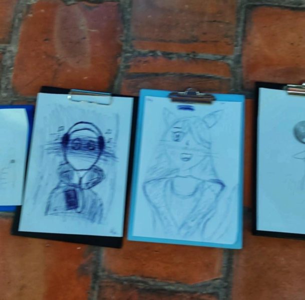 Tekst alternatywny (Alt text): Trzy szkice postaci wykonane na białych kartkach, przypięte do niebieskiej i czarnych podkładek z klipsem, leżące na ceglanej podłodze. Rysunki przedstawiają postacie w słuchawkach oraz z elementami fantastycznymi, wykonane niebieskim długopisem i ołówkiem