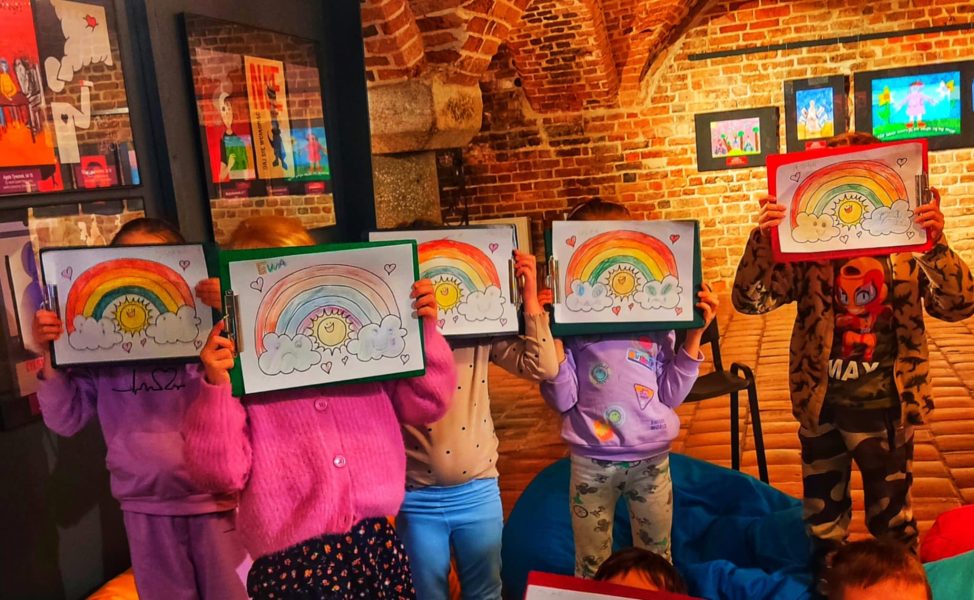 Grupa dzieci w kolorowej sali warsztatowej trzyma przed twarzami tabliczki z pokolorowanymi rysunkami uśmiechniętego słońca i tęczy. W tle widać ceglane łuki oraz galerię oprawionych prac plastycznych