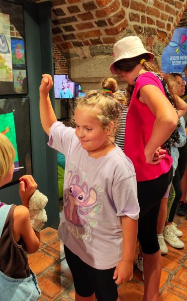 Grupa dzieci w różnym wieku bawi się i gestykuluje w sali o ceglanych sklepieniach. Na pierwszym planie dziewczynka w jasnofioletowej koszulce z uśmiechem unosi rękę, a obok niej stoją inne dzieci oglądające wystawę prac plastycznych