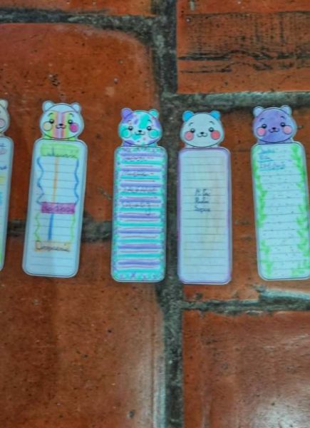 Pięć kolorowych, papierowych zakładek do książek z uśmiechniętymi zwierzątkami na górze, ułożonych na ceglanej podłodze. Zakładki mają miejsce na notatki i są ozdobione pastelowymi kolorami oraz ręcznym pismem