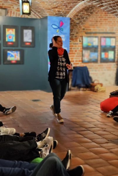 Monika Wojdyło w czarnym swetrze, kraciastej koszuli i pomarańczowym szaliku prowadzi warsztaty dla dzieci w sali o ceglanej architekturze. W tle widoczne są tablice z kolorowymi pracami plastycznymi