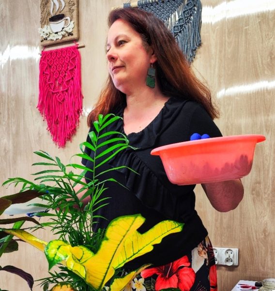 Monika Wojdyło, kobieta z długimi brązowymi włosami, stoi obok roślin doniczkowych (palmy i krotona). Trzyma różową miskę, a w tle na ścianie wiszą kolorowe makramy