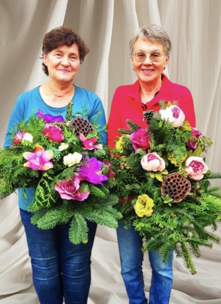 Dwie uśmiechnięte seniorki stoją obok siebie na jasnym tle, prezentując duże, bogate wiązanki nagrobne. Kompozycje wykonane są z gęstego, zielonego igliwia, ozdobione różowymi kwiatami i naturalnym suszem lotosu