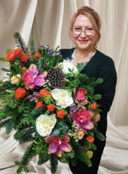 Uśmiechnięta uczestniczka warsztatów trzyma dużą, bogatą wiązankę nagrobną wykonaną z żywego igliwia, białych róż, fioletowych orchidei oraz pomarańczowych lampionów miechunki