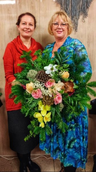 Monika Wojdyło (po lewej, w czerwonym stroju) stoi obok uśmiechniętej kobiety w niebieskiej sukience w kwieciste wzory. Razem trzymają bardzo dużą, bogatą wiązankę nagrobną z zielonego igliwia, różowych kwiatów i żółtych storczyków
