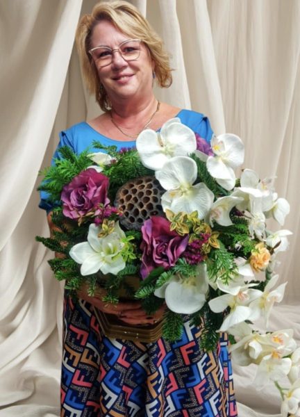 Uśmiechnięta kobieta w okularach i niebieskiej bluzce trzyma bardzo dużą, rozłożystą wiązankę nagrobną. Kompozycja składa się z białych storczyków, ciemnofioletowych róż, zielonego igliwia oraz dużego, dekoracyjnego suszu lotosu w centrum