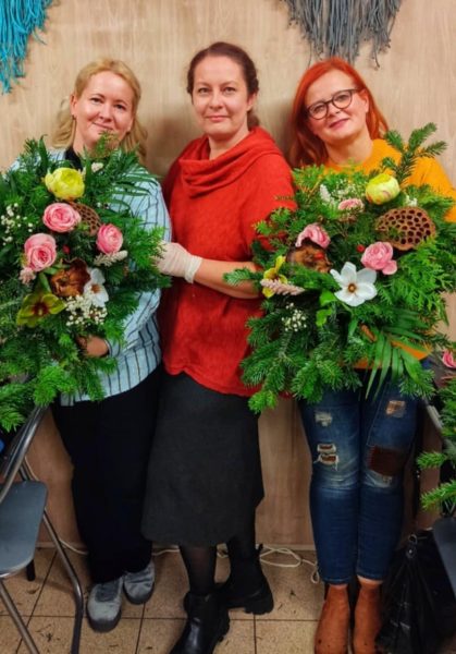 Trzy uśmiechnięte kobiety stoją obok siebie w pracowni, trzymając duże, własnoręcznie wykonane wiązanki nagrobne z żywego igliwia i kolorowych kwiatów. Środkowa postać to Monika Wojdyło w czerwonym stroju