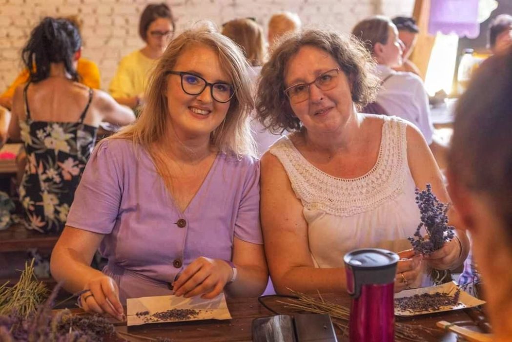 Dwie uśmiechnięte kobiety siedzą obok siebie przy drewnianym stole w rustykalnym wnętrzu, przygotowując suszoną lawendę do woreczków zapachowych