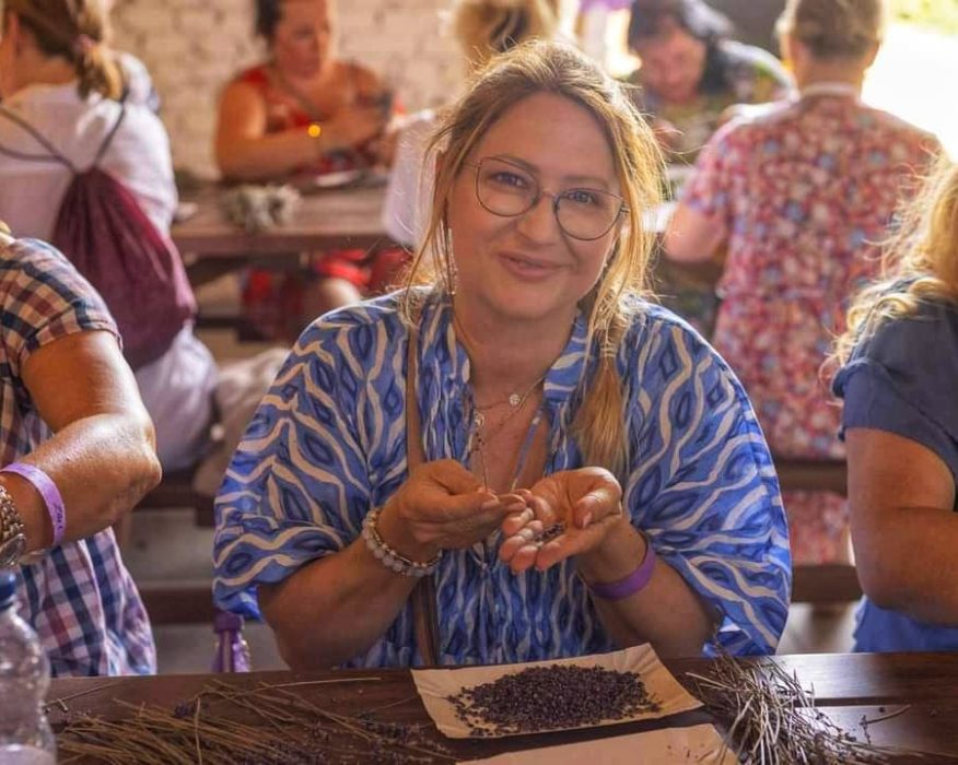 Uśmiechnięta kobieta w okularach i niebieskiej bluzce we wzory siedzi przy drewnianym stole i trzyma w dłoniach ususzone ziarna lawendy podczas warsztatów rękodzieła.