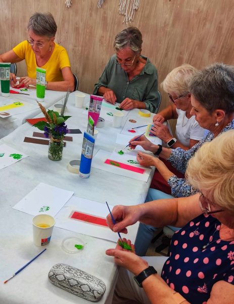 Pięć uśmiechniętych seniorek siedzi przy długim stole przykrytym białym obrusem, intensywnie pracując nad swoimi dziełami. Uczestniczki malują pędzlami na małych formatach, korzystając z kolorowych farb w tubkach (zielonych, różowych, niebieskich)