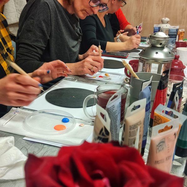 Trzy kobiety siedzące przy wspólnym stole, skupione na malowaniu mandali kropkowych na okrągłych, czarnych podkładach. Na stole znajdują się tubki z farbami akrylowymi, pędzle, naczynia z wodą oraz zapalona srebrna latarenka. Na pierwszym planie widoczny jest rozmyty fragment czerwonej róży