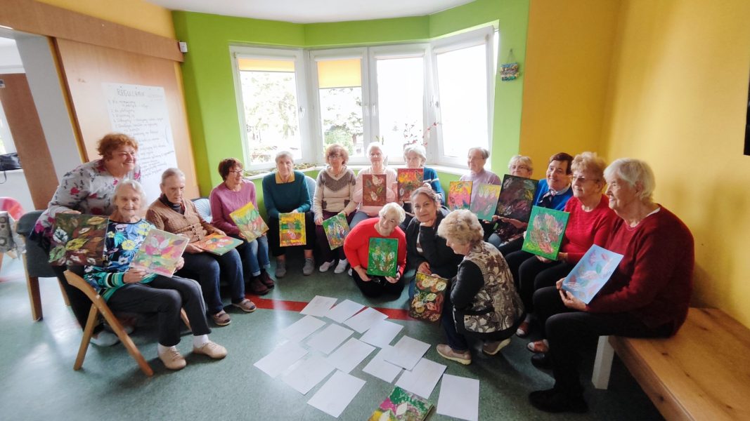 Grupowe zdjęcie kilkunastu seniorek siedzących w półkolu w jasnym, zielono-żółtym pokoju z dużym oknem. Każda z pań trzyma na kolanach własnoręcznie namalowany obraz przedstawiający kolorowe kompozycje z jesiennych liści. Na podłodze przed nimi leżą kolejne arkusze papieru i obrazy. Kobiety uśmiechają się do obiektywu, prezentując efekty swojej pracy