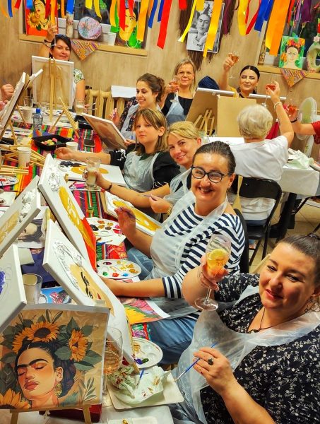 Grupa kobiet podczas warsztatów malowania przy winie, tworzących obraz Fridy Kahlo na płótnach, z kieliszkami wina, farbami i kolorowymi dekoracjami artystycznymi w tle Warsztatowni