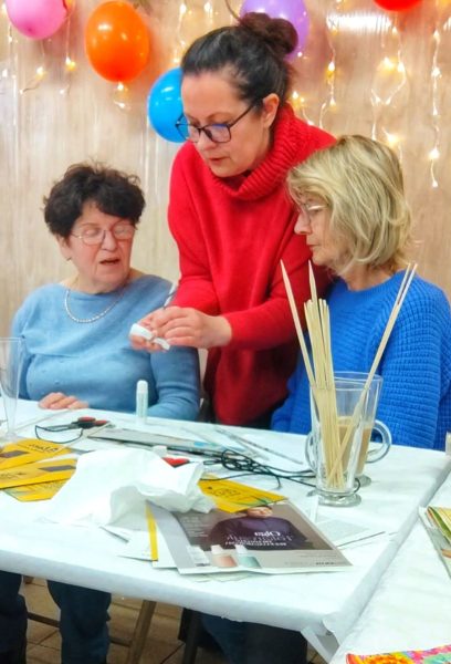 Monika Wojdyło, ubrana w czerwony golf, pochyla się nad stołem warsztatowym, pomagając dwóm uczestniczkom w precyzyjnym skręcaniu rurek z wikliny papierowej. Na białym obrusie leżą materiały edukacyjne, klej, nożyczki oraz wysoka szklanka z długimi, jasnymi patyczkami do skręcania papieru. W tle widać dekoracyjne lampki i kolorowe balony, tworzące świąteczną atmosferę zajęć