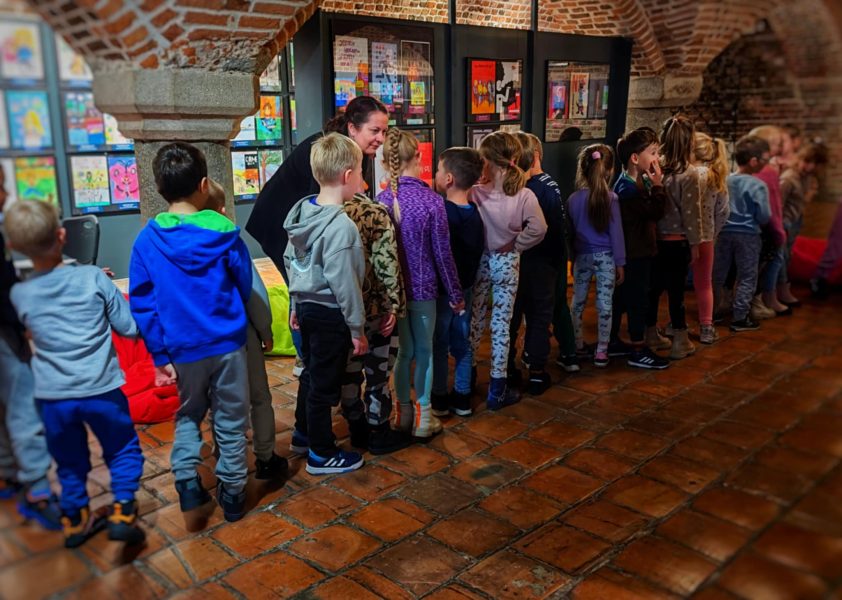 Grupa dzieci ustawiona w długim rzędzie na środku sali o ceglanej architekturze. Prowadząca Monika Wojdyło w czarnym stroju pochyla się nad jednym z chłopców na początku kolejki, rozmawiając z nim. W tle widać wystawę kolorowych prac plastycznych