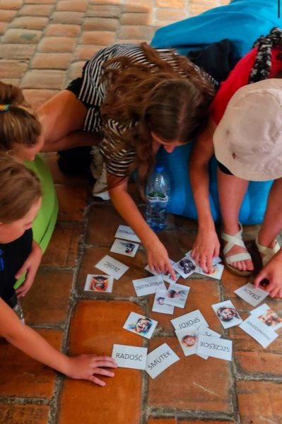 Grupa dzieci siedzących na kolorowych pufach i na ceglanej podłodze, pochylona nad rozłożonymi kartami z obrazkami i napisami określającymi emocje, takimi jak: Radość, Smutek, Złość, Strach, Spokój, Szczęście