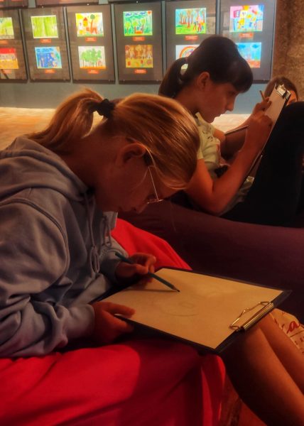 Dwie dziewczynki siedzą na wygodnych pufach w nastrojowo oświetlonej sali z wystawą prac plastycznych w tle. Uczestniczki są skupione na rysowaniu ołówkiem na kartkach umieszczonych na sztywnych podkładkach z klipsem