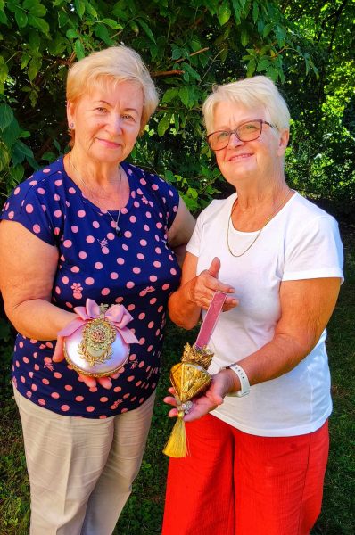 Dwie uśmiechnięte kobiety w wieku dojrzałym stoją w ogrodzie na tle zielonych krzewów, prezentując swoje prace rękodzielnicze. Jedna trzyma różową, bogato zdobioną bombkę z atłasową kokardą, druga – złotą dekorację w kształcie serca z chwostem