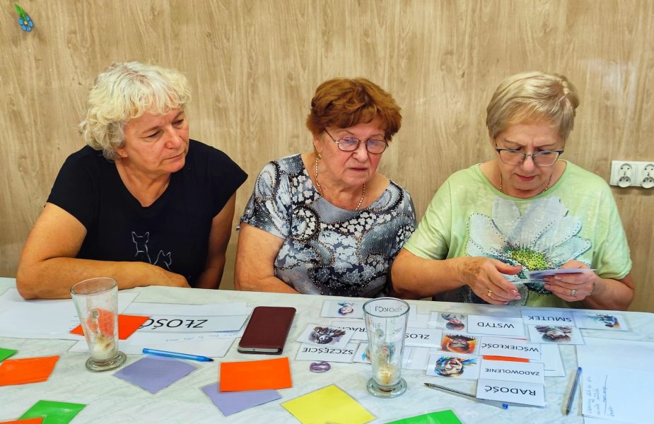 Trzy starsze kobiety siedzą przy stole, skupione na analizowaniu kart z nazwami emocji, takimi jak Radość, Złość, Smutek i Wstyd, podczas warsztatów psychologicznych