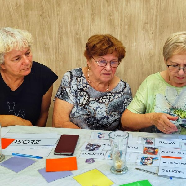 Trzy starsze kobiety siedzą przy stole, skupione na analizowaniu kart z nazwami emocji, takimi jak Radość, Złość, Smutek i Wstyd, podczas warsztatów psychologicznych