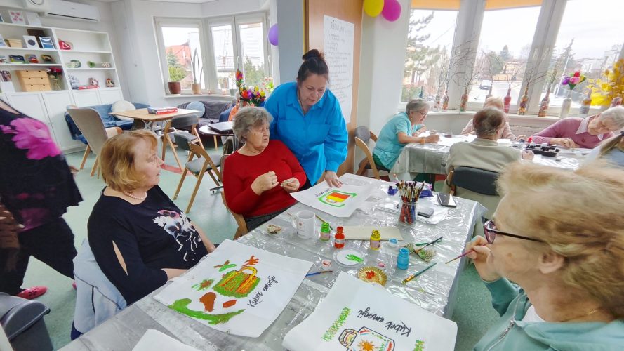 Grupa seniorów podczas warsztatów „Emocje w kolorach” w jasnej sali, przy stołach z farbami, pędzlami i pracami plastycznymi, z prowadzącą wspierającą uczestników w twórczym działaniu