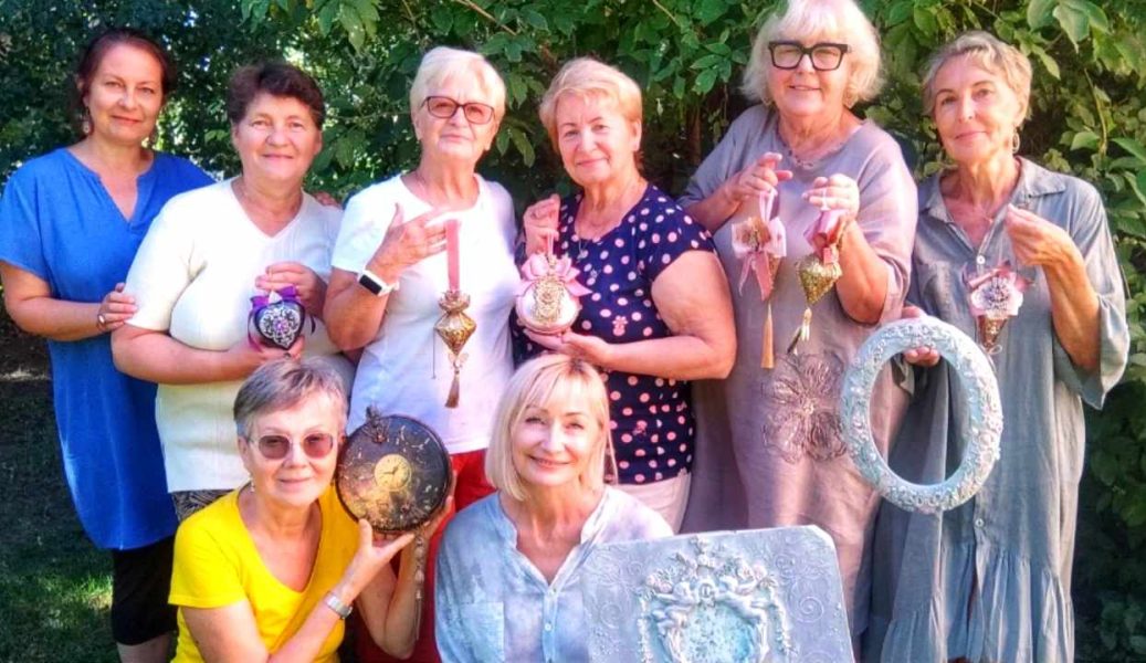 Grupa uśmiechniętych seniorek stoi w ogrodzie wraz z Moniką Wojdyło (po lewej, w niebieskiej bluzce). Kobiety dumnie prezentują wykonane w technice decoupage przedmioty: ozdobne zegary, bombki, serca oraz bogato zdobioną ramę