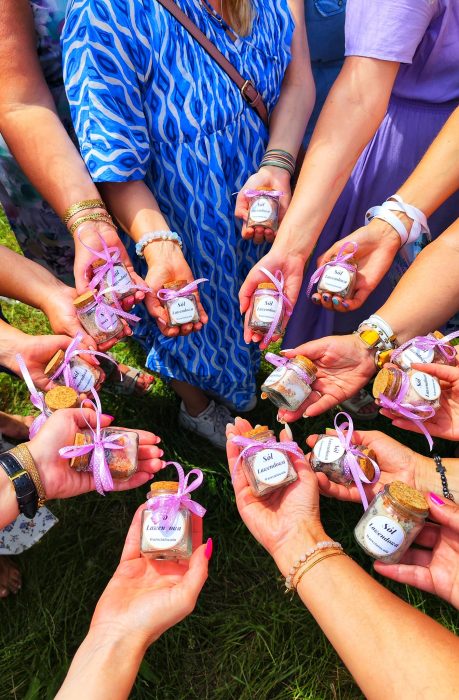 Widok z góry na dłonie wielu uczestniczek ułożone w kręgu, z których każda trzyma mały szklany słoiczek z solą lawendową Warsztatowni, ozdobiony różową wstążką