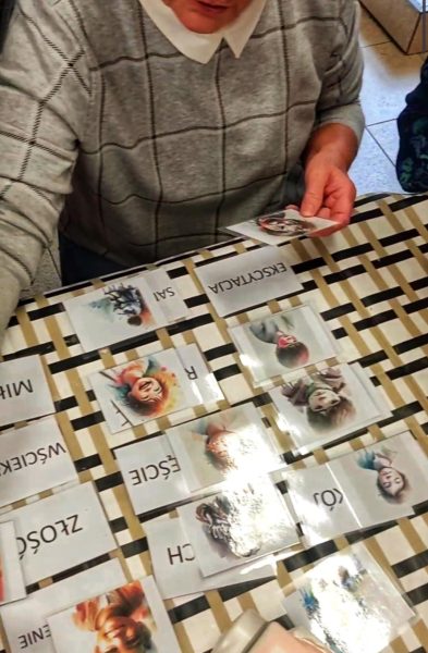 Seniorka podczas warsztatów terapeutycznych wybiera ilustracje przedstawiające różne twarze i sytuacje, dopasowując je do kart z nazwami emocji, takimi jak „Ekscytacja”, „Złość” czy „Lęk”