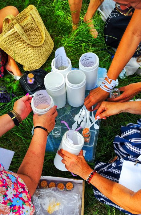 Widok z góry na dłonie kilku osób, które białymi łyżeczkami nabierają różne rodzaje soli (m.in. himalajską) z dużych białych pojemników do małych szklanych słoiczków