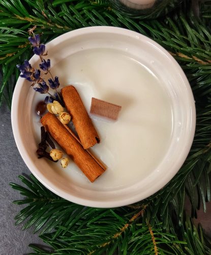 Widok z góry na białą świecę sojową w ceramicznym naczyniu, udekorowaną laskami cynamonu, suszoną lawendą, goździkami i pączkami kwiatów. Świeca ma drewniany knot i spoczywa na podkładzie z zielonych gałązek iglastych