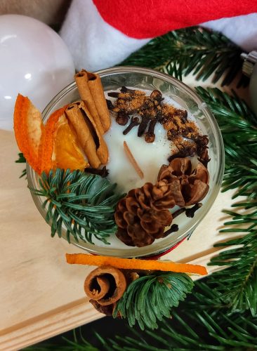 Widok z góry na okrągłą świecę sojową w szklanym naczyniu, udekorowaną laskami cynamonu, goździkami, suszoną pomarańczą oraz szyszkami i gałązkami iglastymi. Świeca z drewnianym knotem jest dodatkowo posypana mielonym cynamonem. W tle widoczna czerwona czapka mikołajkowa oraz biała i srebrna bombka