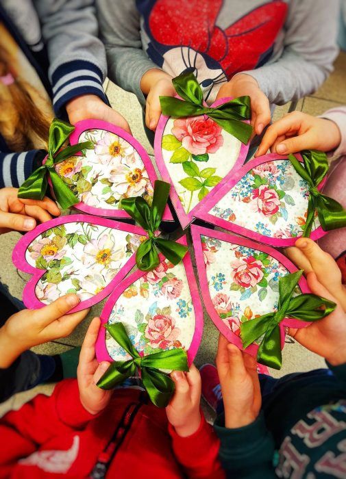 Grupa dzieci trzymająca w dłoniach własnoręcznie ozdobione, duże serca z motywem róż, wykończone różową ramką i zieloną satynową kokardą. Serca ułożone w kształt kwiatu