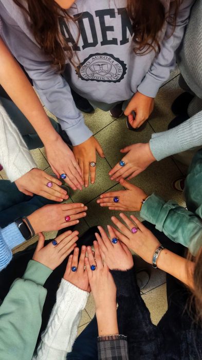 Grupa dzieci trzyma dłonie w kole, prezentując kolorowe, własnoręcznie wykonane pierścionki z żywicy w różnych odcieniach: różowym, niebieskim i fioletowym