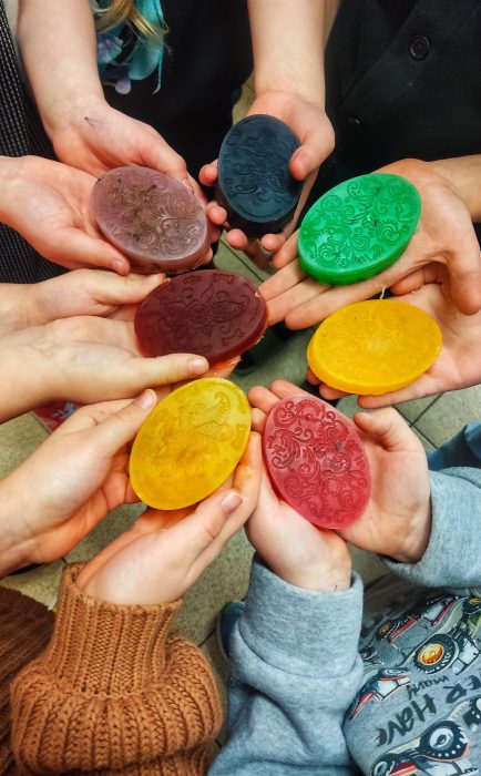 Grupa dzieci trzyma dłonie w kole, prezentując kolorowe, własnoręcznie wykonane mydełka glicerynowe z ozdobnymi wzorami w kolorach: zielonym, żółtym, czerwonym, fioletowym i czarnym