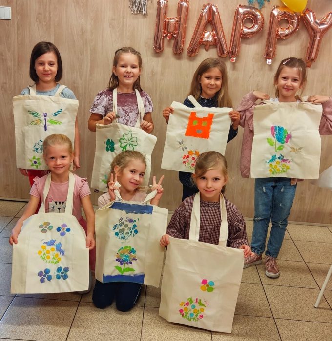 Siedem uśmiechniętych dziewczynek prezentuje białe torby bawełniane z kolorowymi nadrukami (motyle, kwiaty, koty) wykonanymi podczas warsztatów. W tle widoczny różowy napis balonowy „HAPPY”